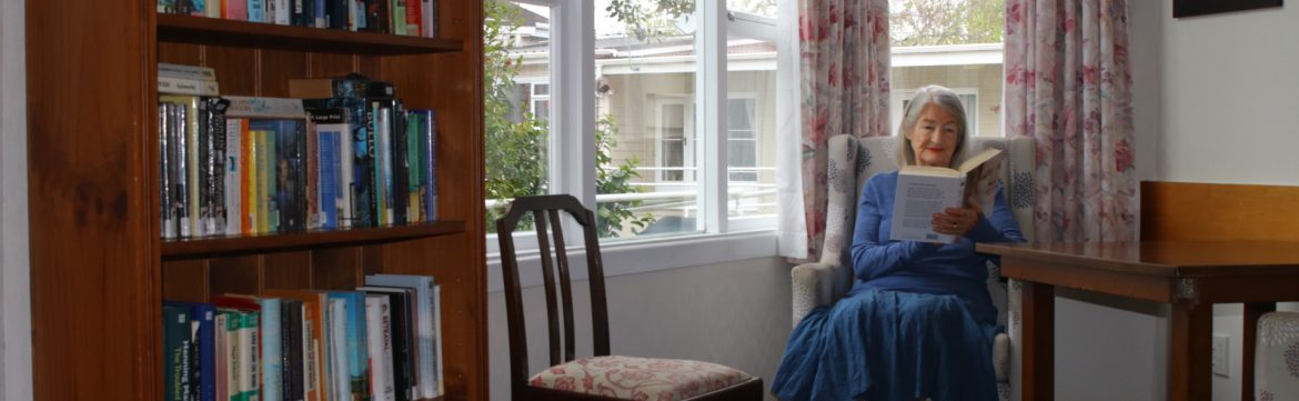 Sunrise Healthcare Ltd elderly woman sitting in a chair reading next to a window with a bookshelf in view.