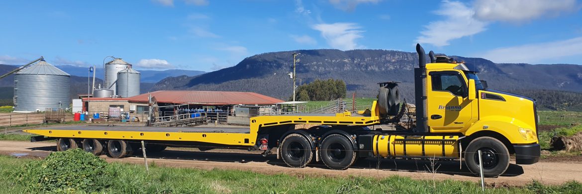 Brianna Tilt Trays & Towing Pty Ltd. yellow semi with towing trailer on a road with a mountain behind.
